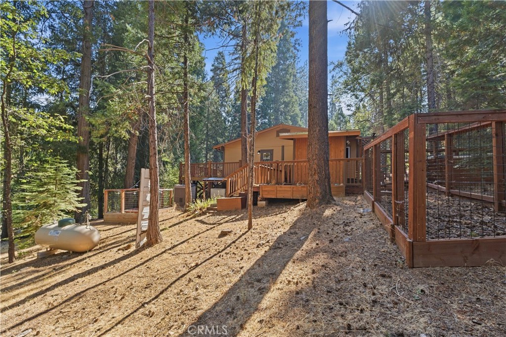 Wood-sided home among tall trees, with a raised deck, fenced yard, and propane tank.