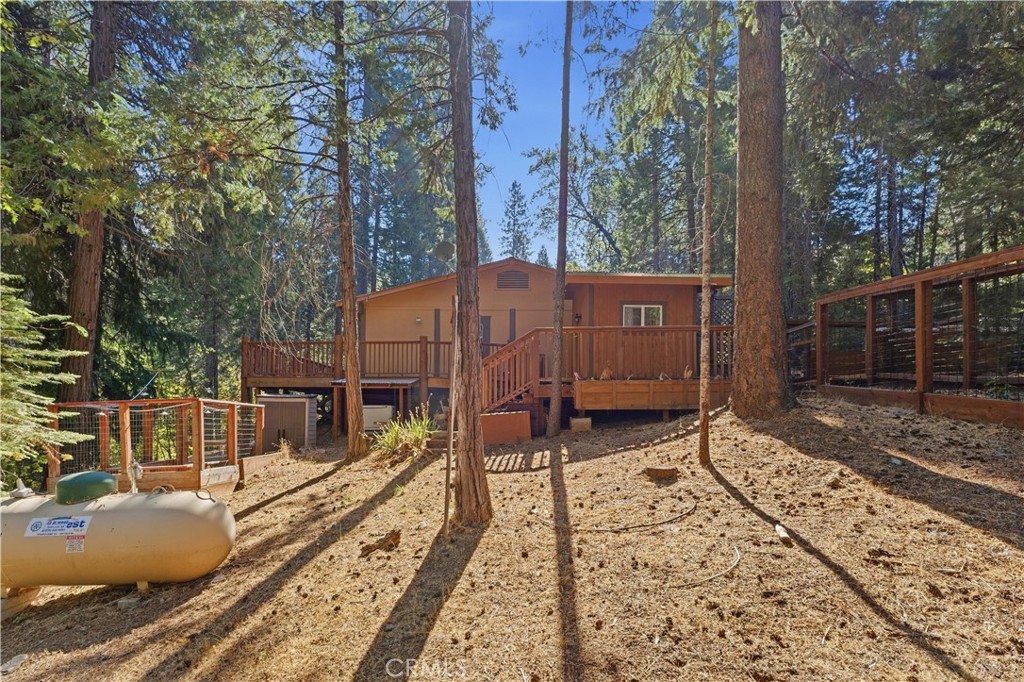 Wood-sided house with a raised deck among tall conifers on a sloping forested lot.
