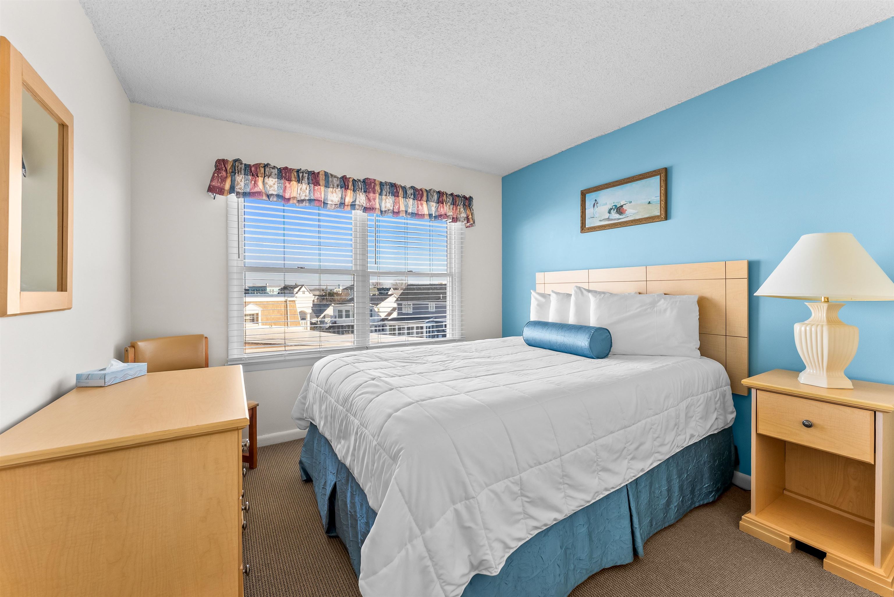 Bedroom with a blue accent wall, neatly made bed, and window overlooking neighboring homes.