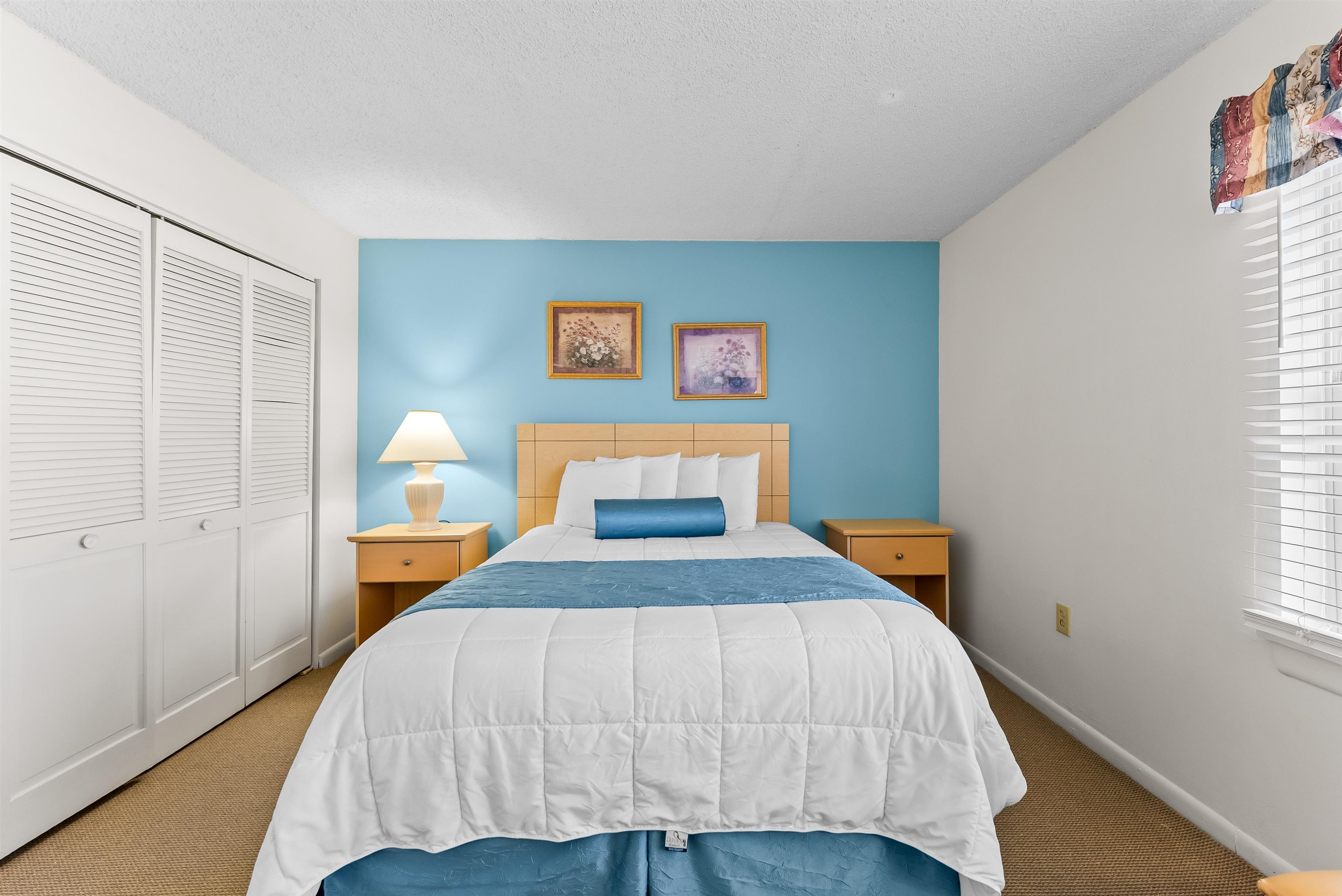 Furnished bedroom with a white bed, blue accents, turquoise wall, and louvered closet doors.