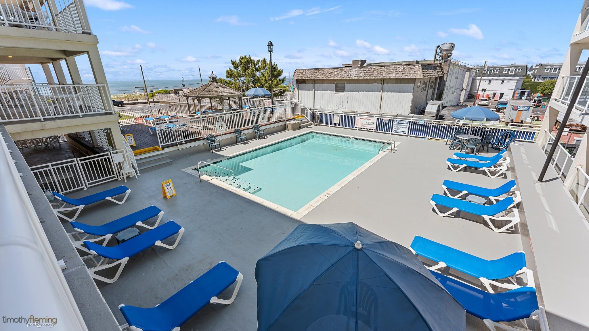 Outdoor pool with blue lounge chairs and umbrellas, with the ocean visible beyond the deck.