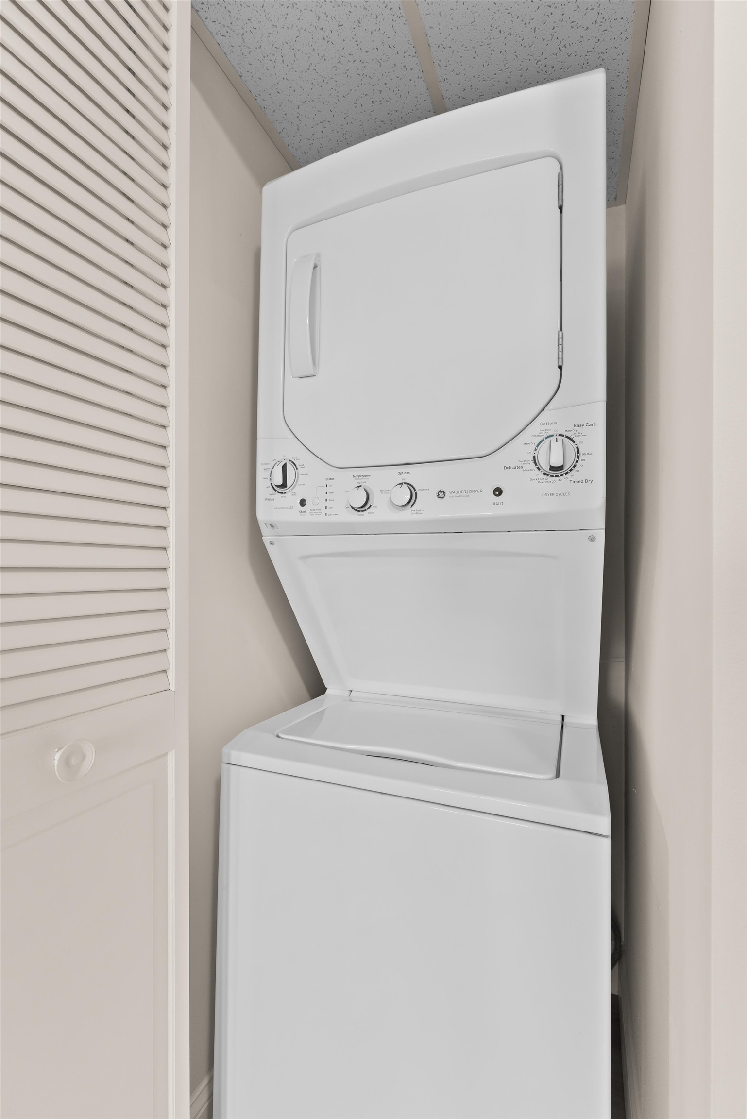 Stacked white washer and dryer in a narrow laundry closet beside louvered doors.