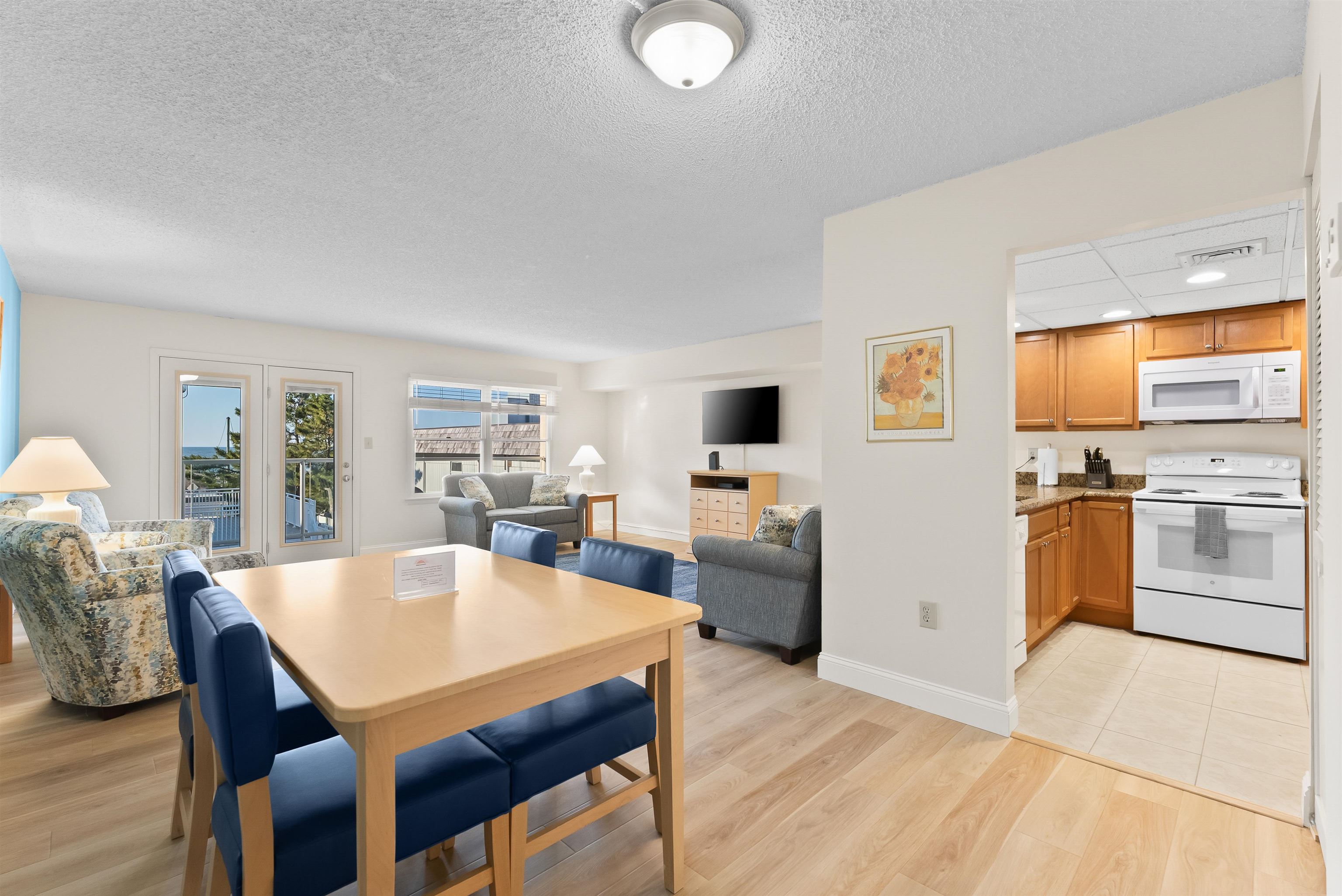 Furnished condo dining and living area with open wood-cabinet kitchen and balcony doors showing water.