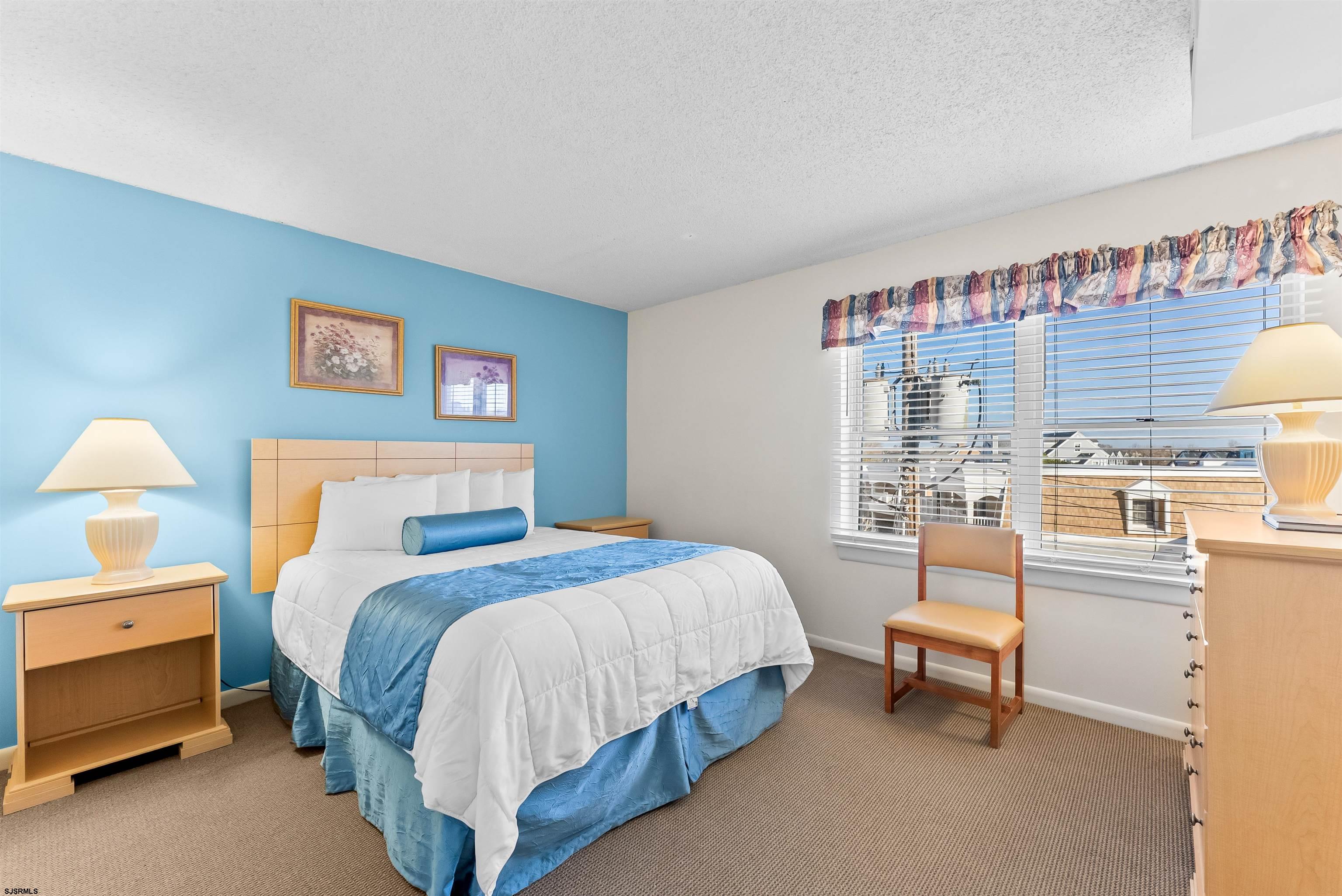 Furnished bedroom with a blue accent wall, white bed, and large window overlooking nearby houses.