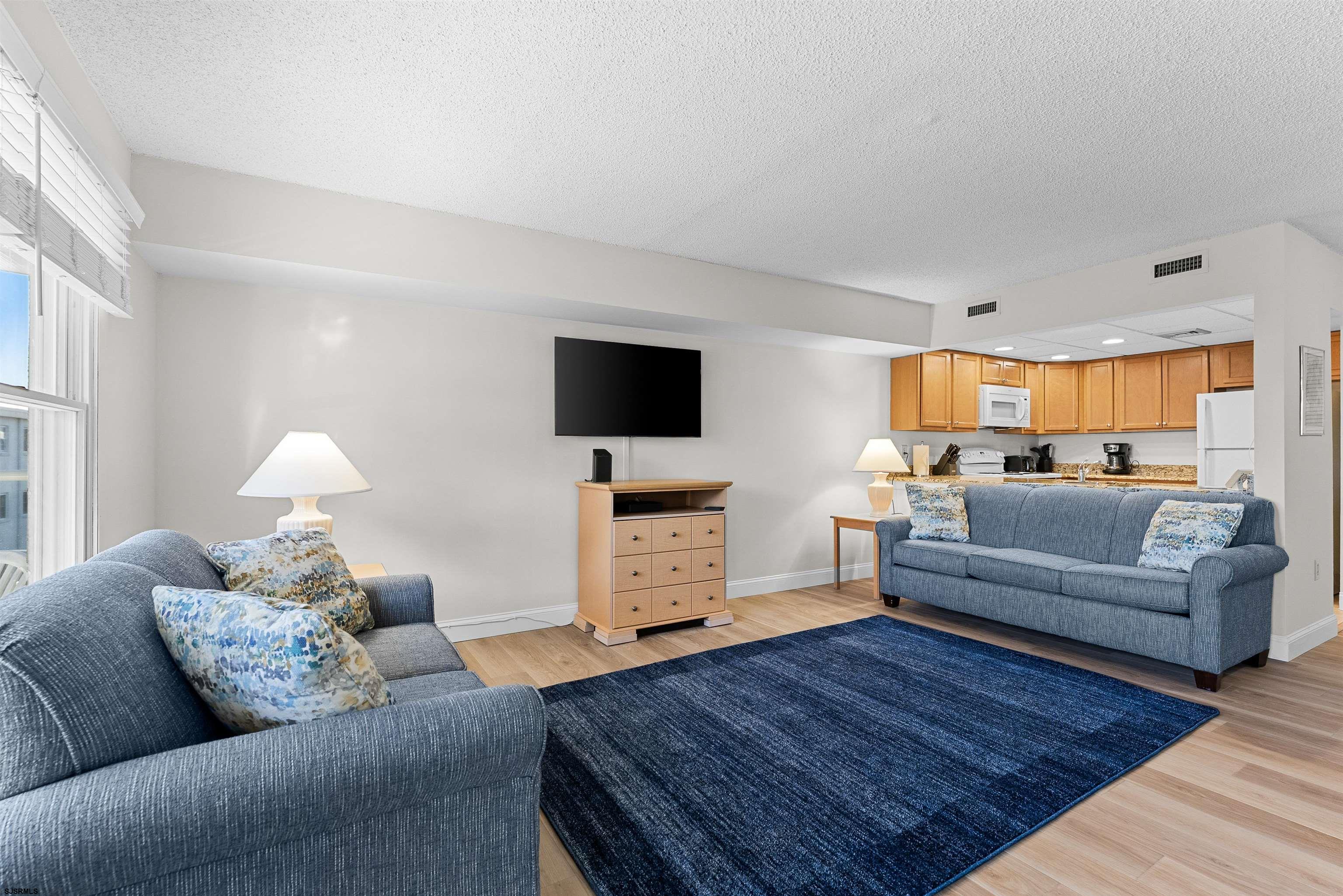 Living room with blue sofas, a dark blue rug, and an open kitchen with wood cabinets.