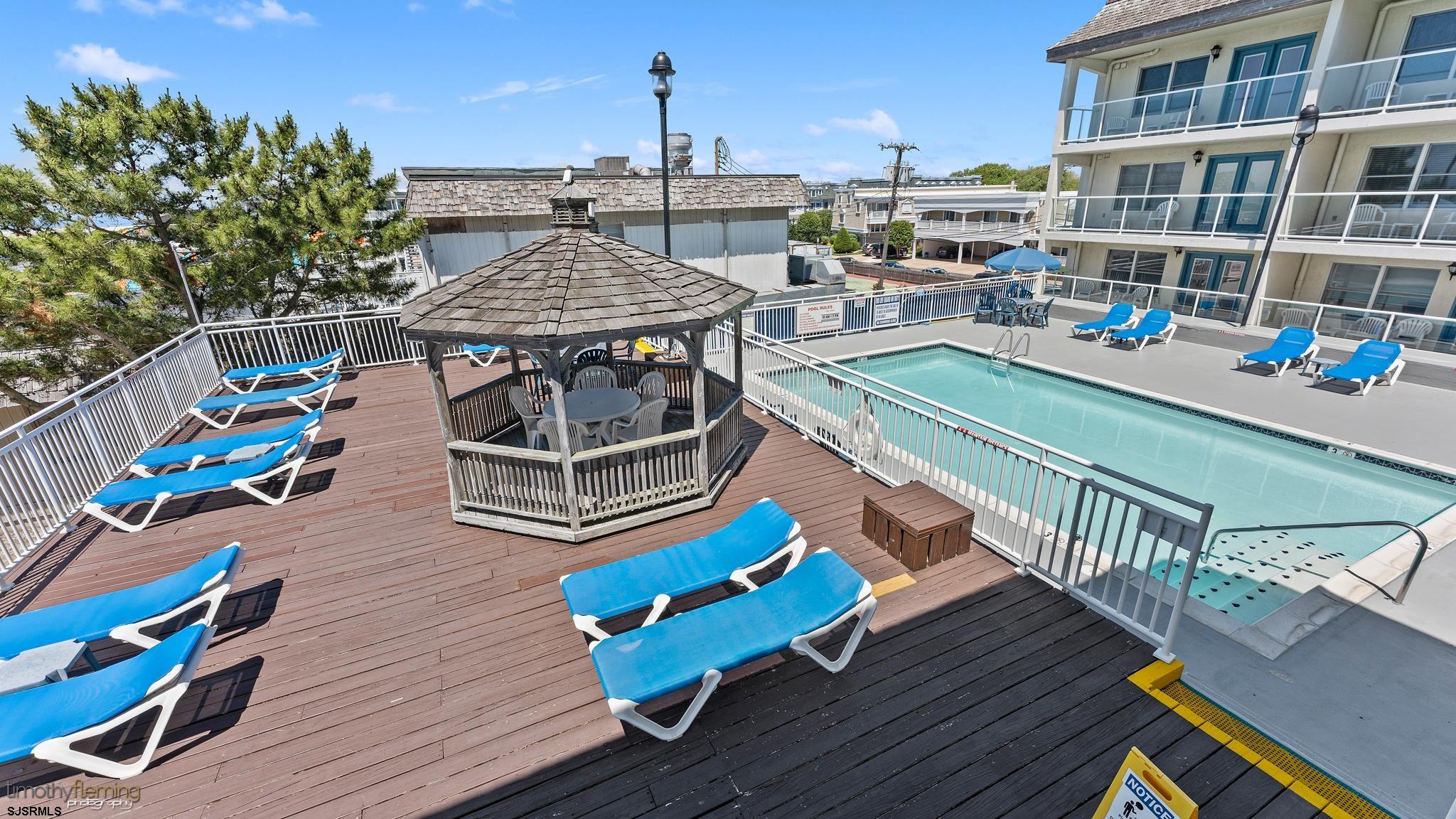 Condominium pool deck with blue lounge chairs, a wooden gazebo, and the building alongside.