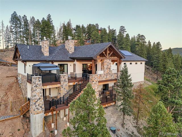 Mountain home with stone columns, timber gables, balconies, and rooftop solar panels on a wooded hillside.