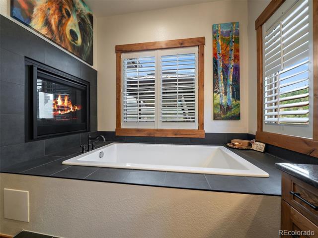 Bathroom with a white soaking tub beside a lit fireplace and shuttered windows.