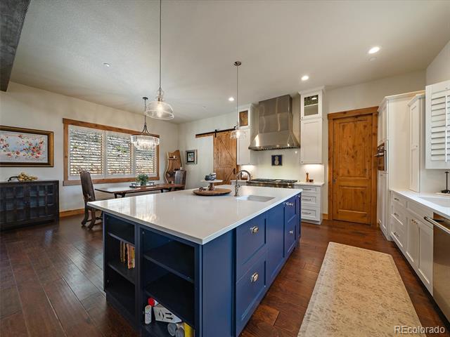 Kitchen with a large blue island, white counters, stainless-steel range hood, and dining area by shuttered windows.