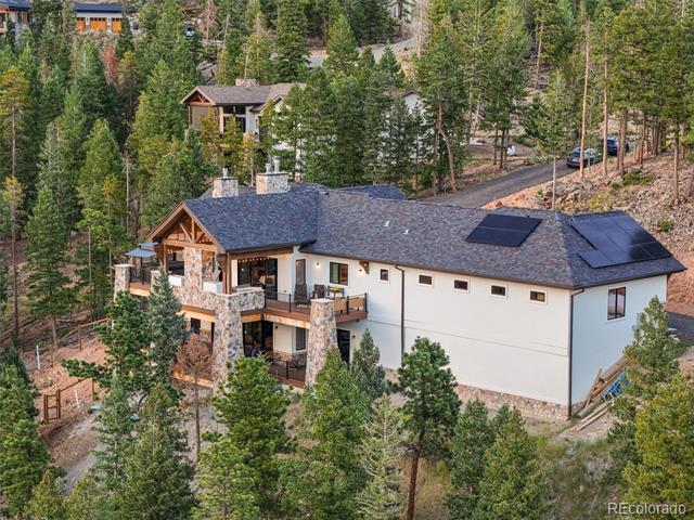 Hillside home surrounded by evergreens, with stone columns, a covered deck, and rooftop solar panels.