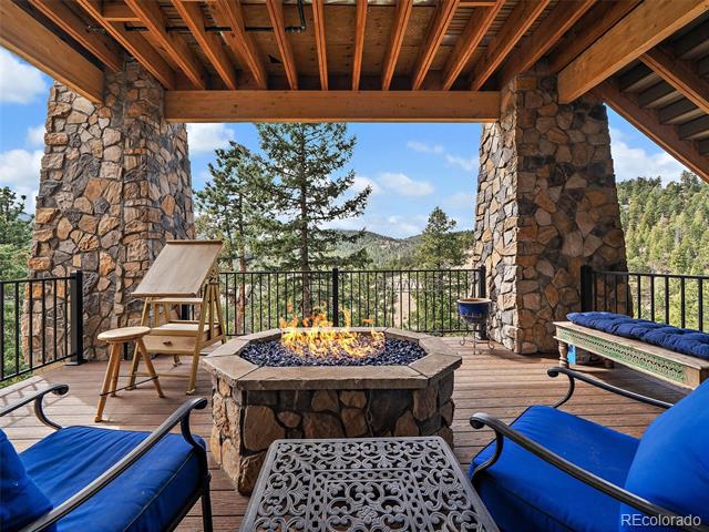 Covered deck with a stone fire pit and blue lounge chairs overlooking wooded mountains.