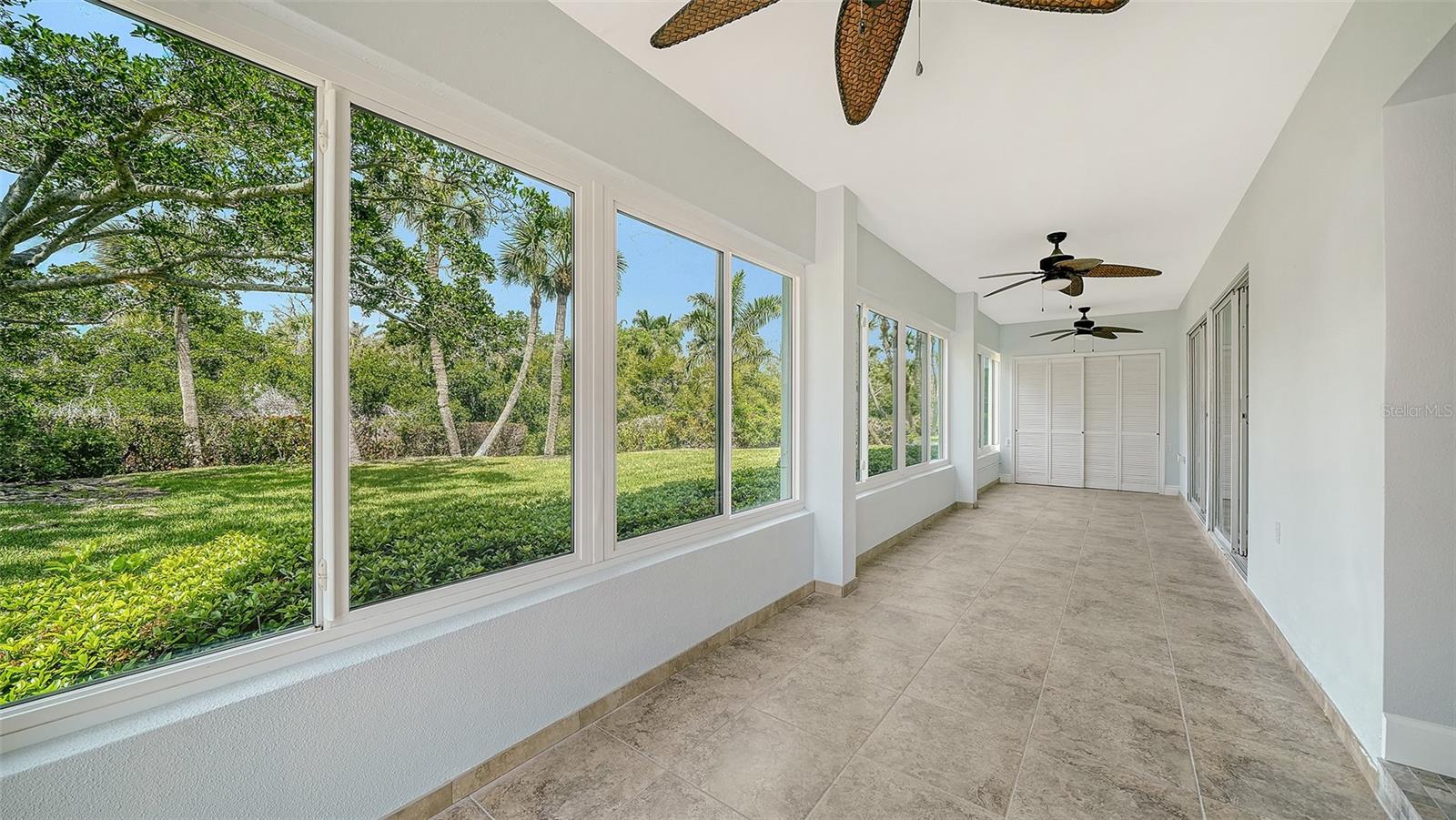 Enclosed lanai with wide garden-facing windows, beige tile flooring, and ceiling fans.
