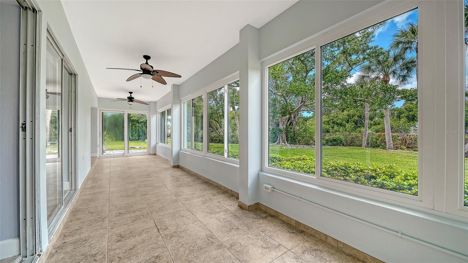 Vacant sunroom with tiled floors, ceiling fans, and large windows overlooking a leafy lawn.