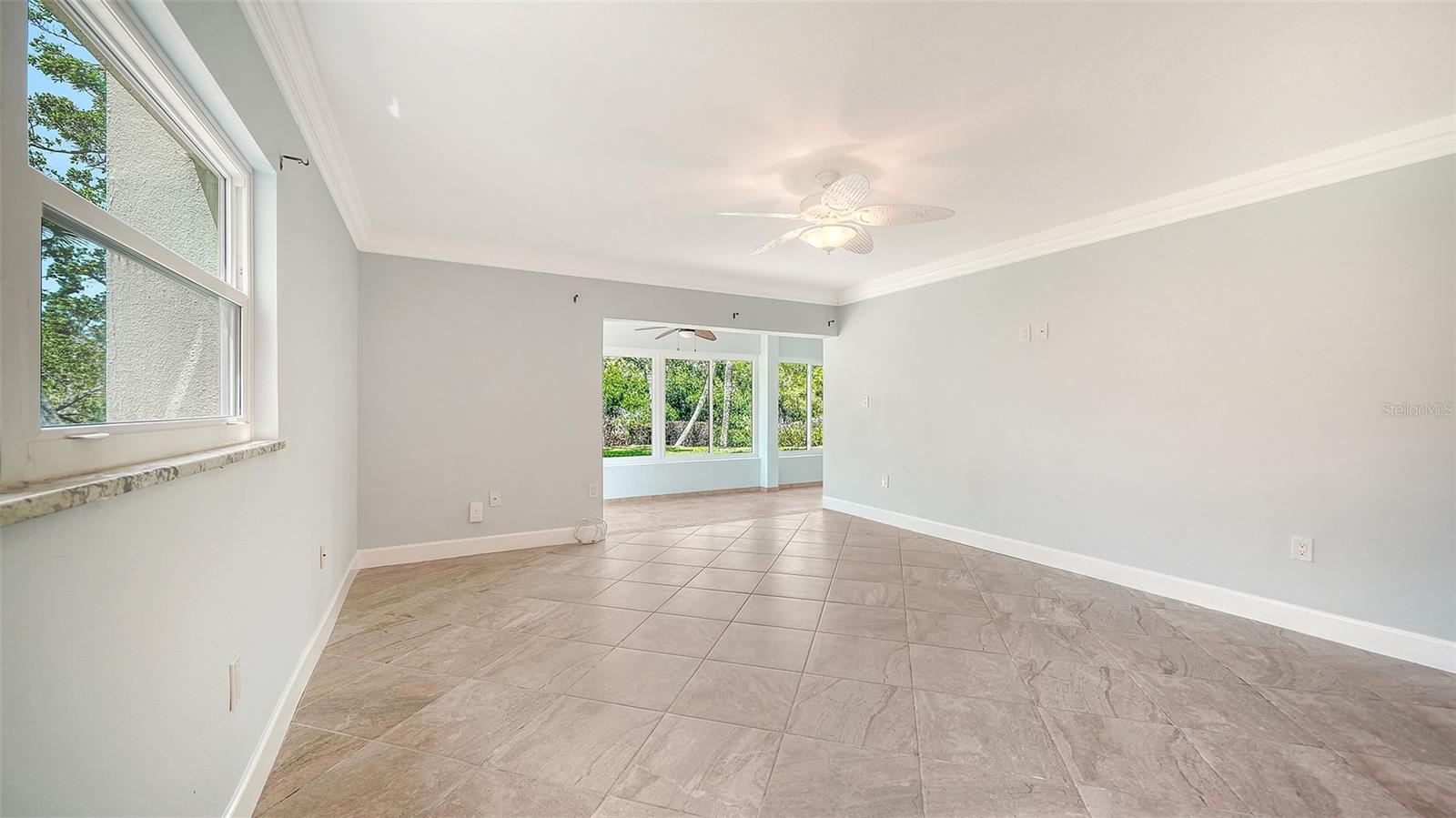 Empty living room with large tile floors, a ceiling fan, and an opening to a bright windowed room.