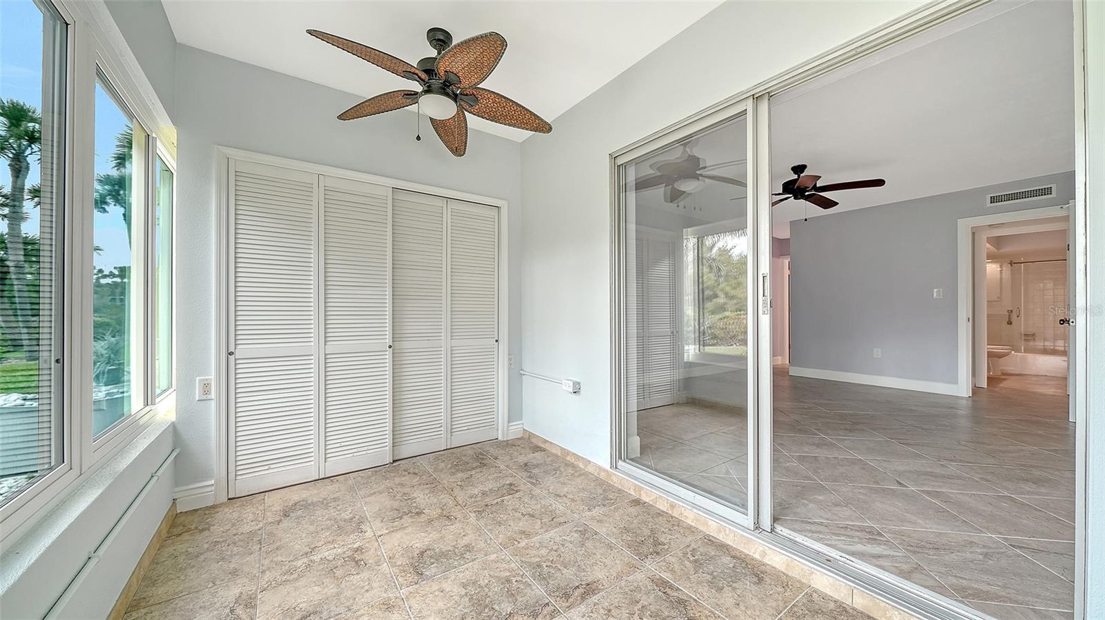 Enclosed sunroom with tiled floors, louvered closet doors, and windows overlooking greenery.
