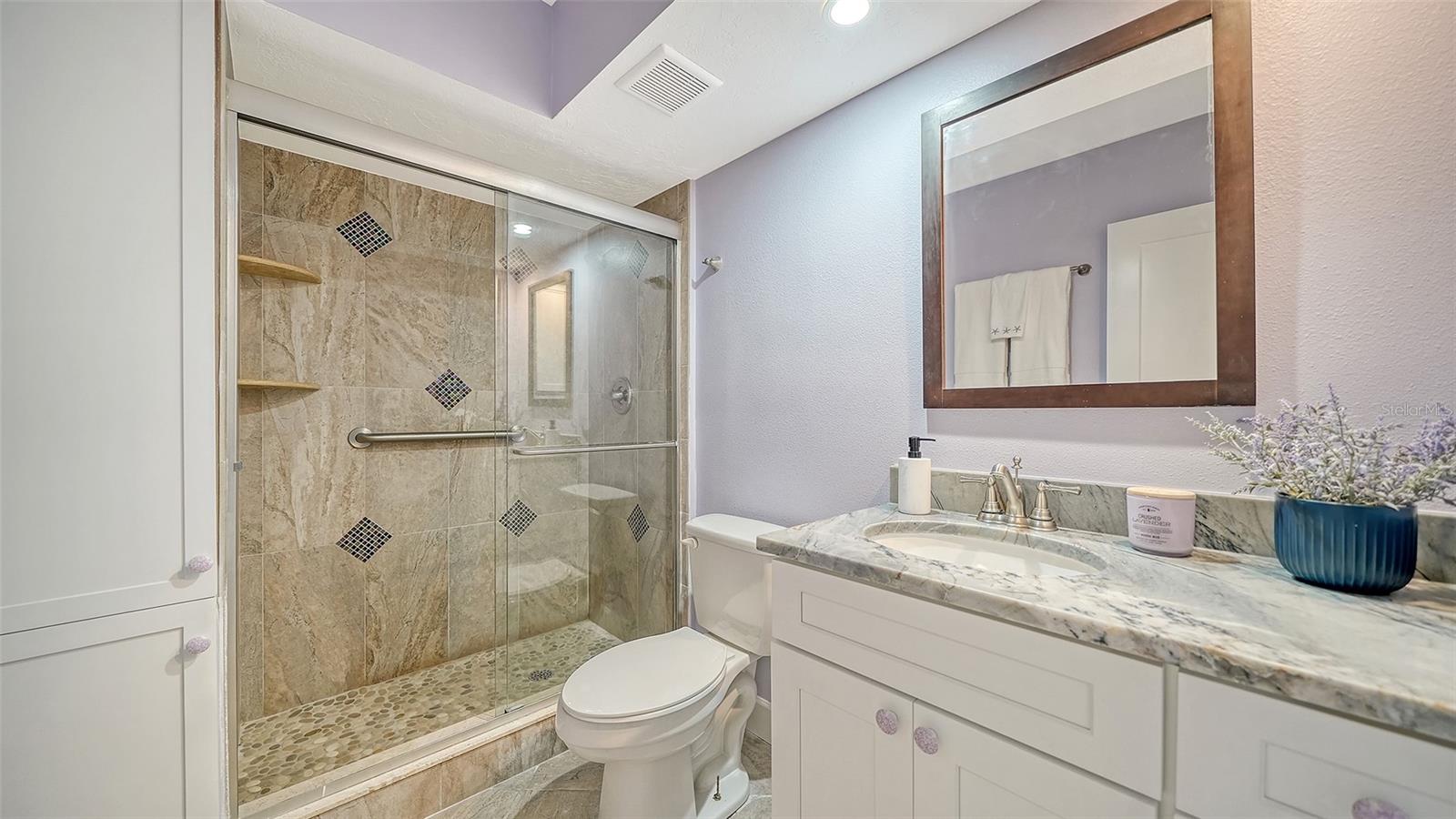 Bathroom with a stone-tiled glass shower, pebble floor, and white vanity with marble countertop.