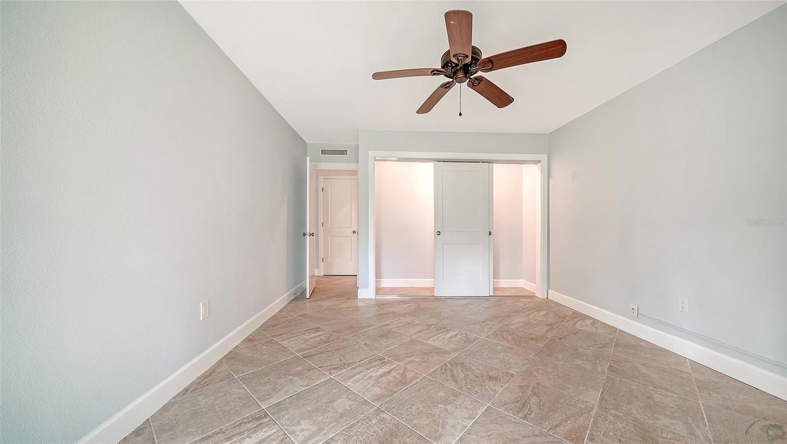 Empty bedroom with tiled floors, a wood-blade ceiling fan, and sliding closet doors.