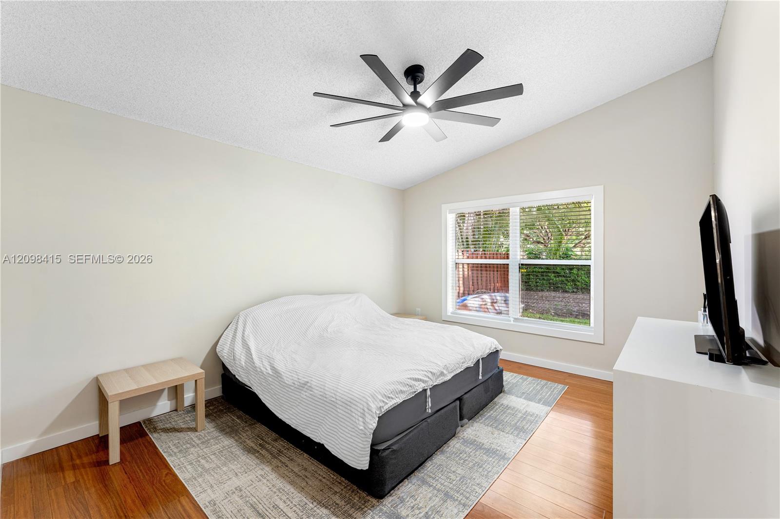 Bedroom with a white-covered bed, large window with blinds, and dark ceiling fan.