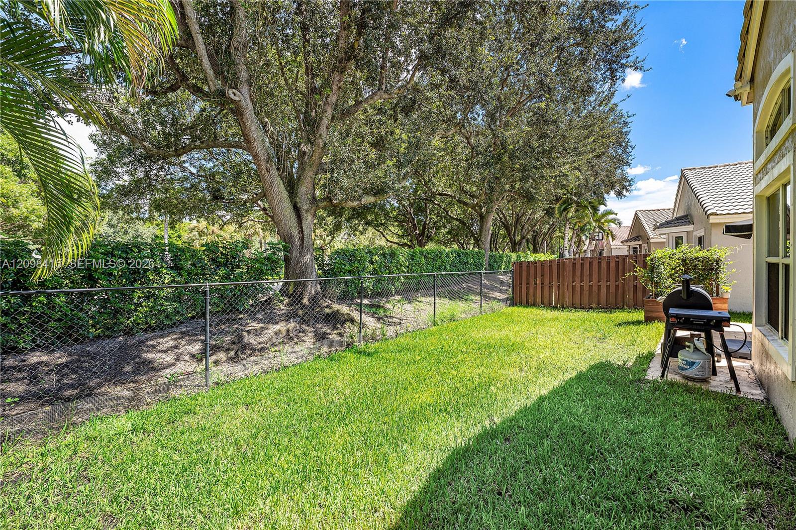 Fenced backyard with a green lawn, mature shade trees, and a barbecue grill beside the home.