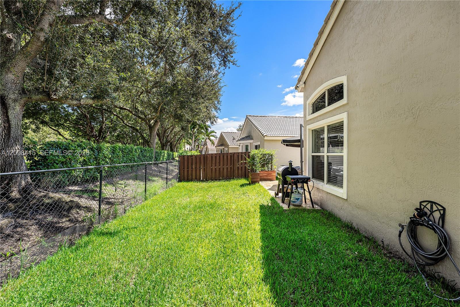 Side yard with a broad lawn, mature trees, chain-link fence, and barbecue grill beside a beige home.