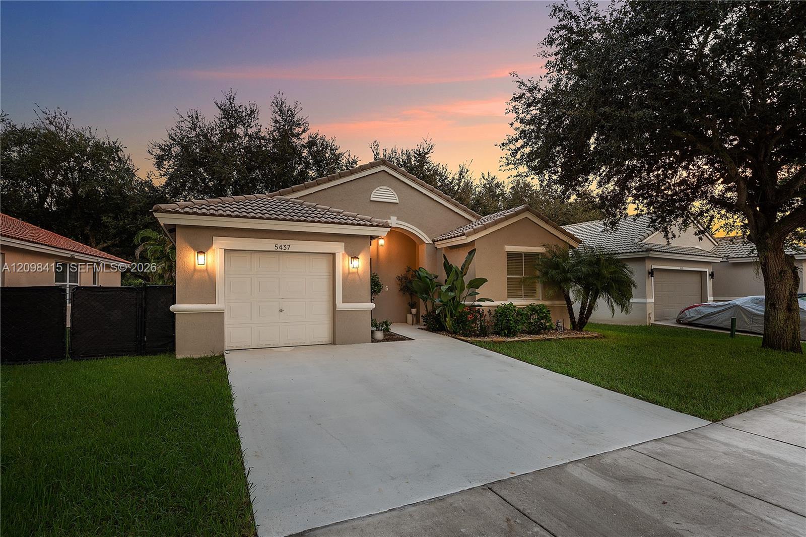 Single-story stucco home with a tiled roof, attached garage, and tropical landscaping at dusk.