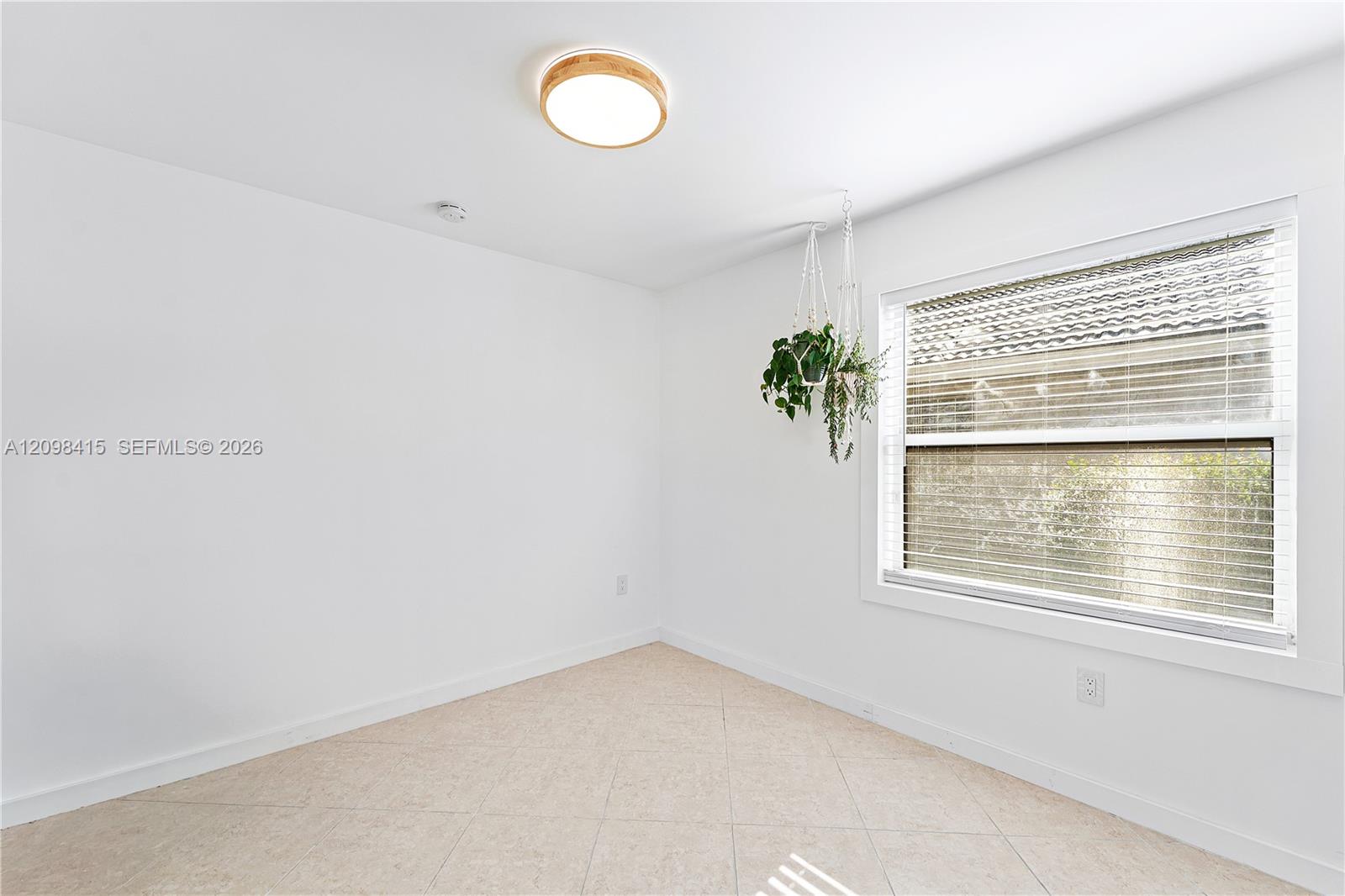 Empty white room with beige tile flooring, window blinds, and hanging plants.