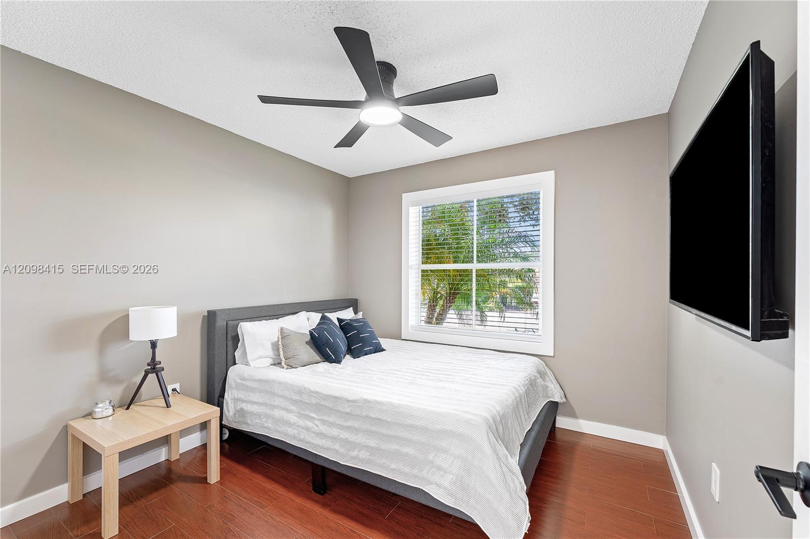 Bedroom with a gray bed, navy pillows, palm-view window, ceiling fan, and wall-mounted television.