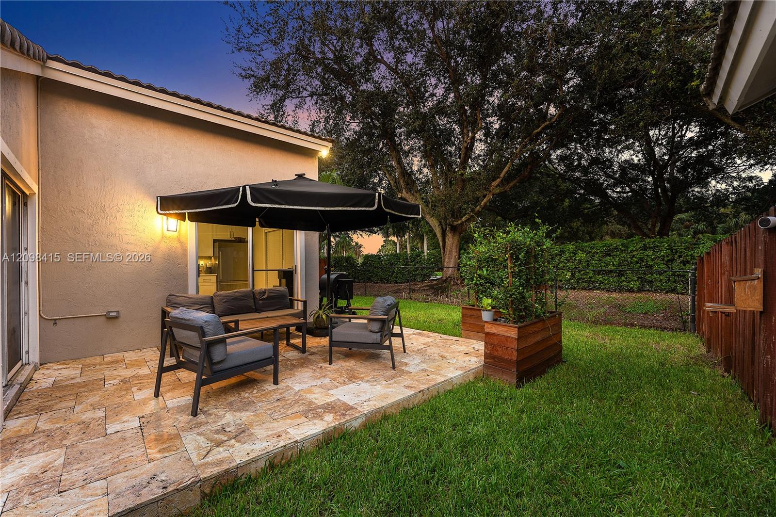 Backyard stone patio with umbrella-shaded seating, a raised planter, and lawn at dusk.