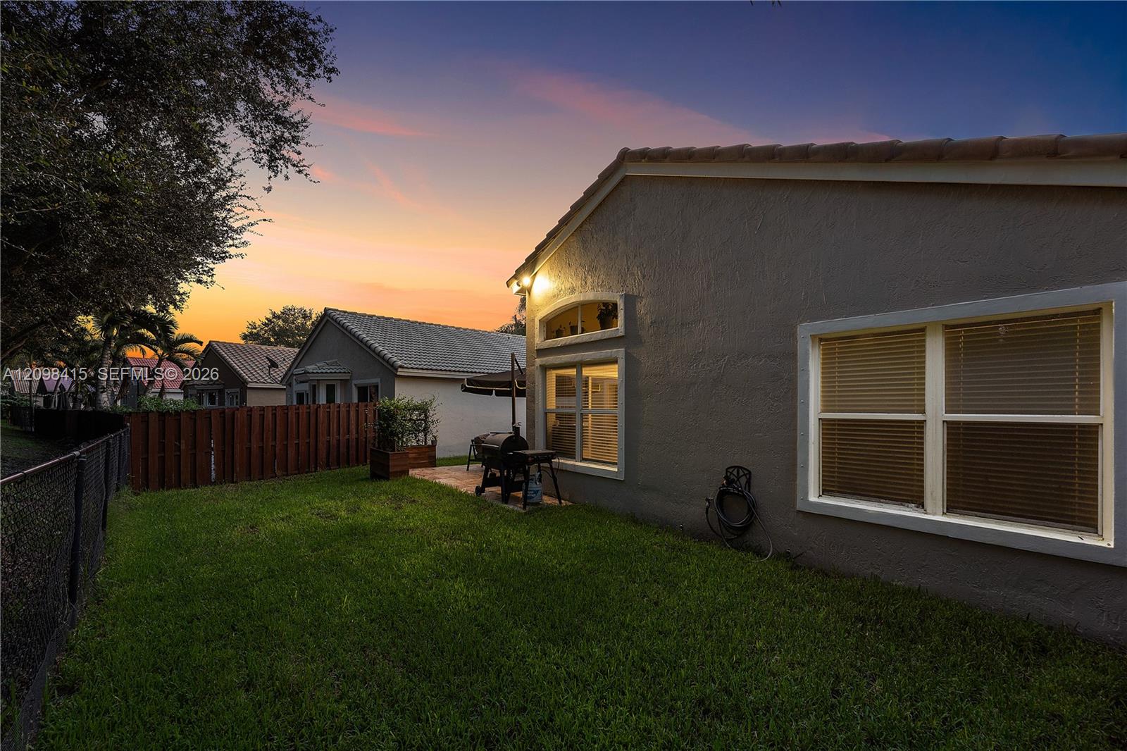 Gray stucco home and grassy backyard at dusk, with a small patio and grill.