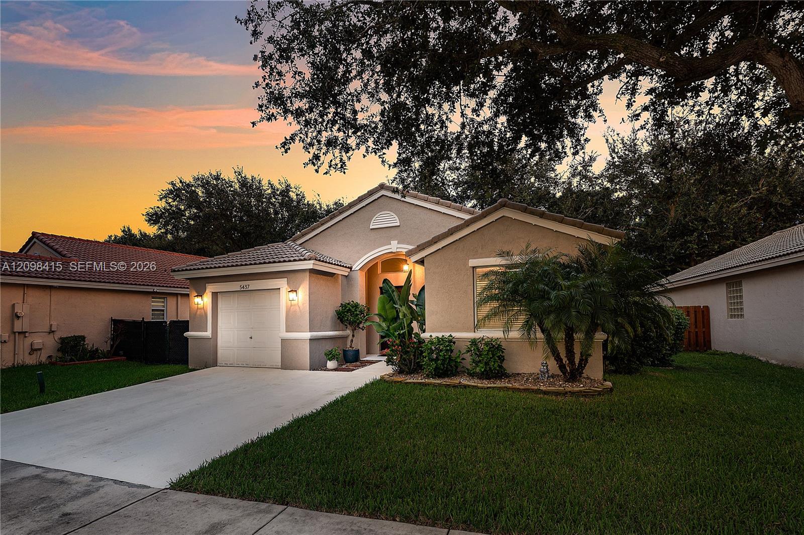 Single-story stucco home with an attached garage, tropical landscaping, and a broad driveway at dusk.