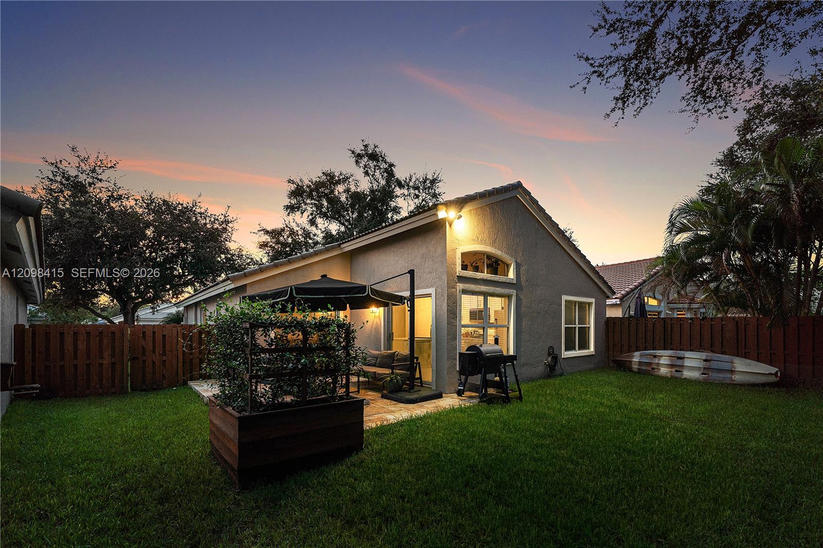 Home’s rear exterior at dusk, with a fenced lawn, covered patio seating, and warmly lit windows.