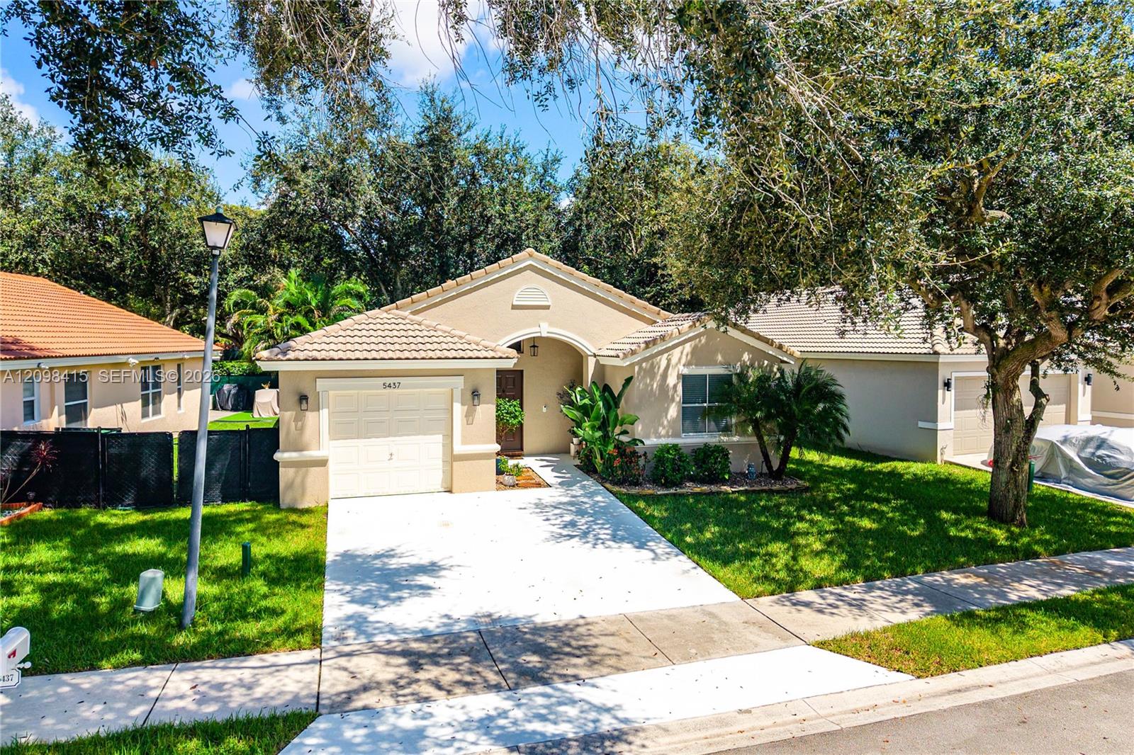 Single-story beige home with a tiled roof, attached garage, and tree-shaded lawn.