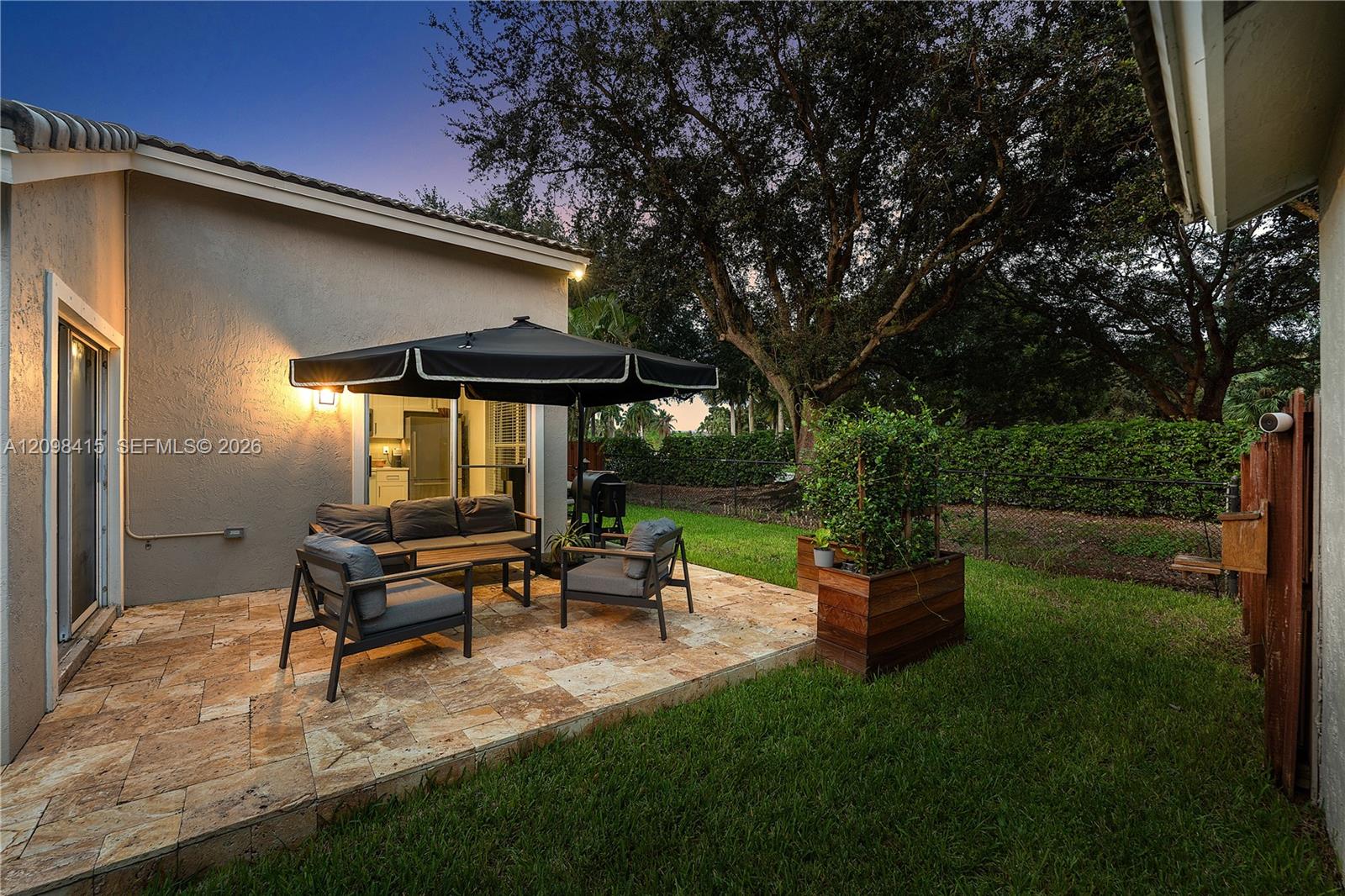 Backyard patio with stone tiles, umbrella-covered seating, and a lawn bordered by hedges and mature trees.