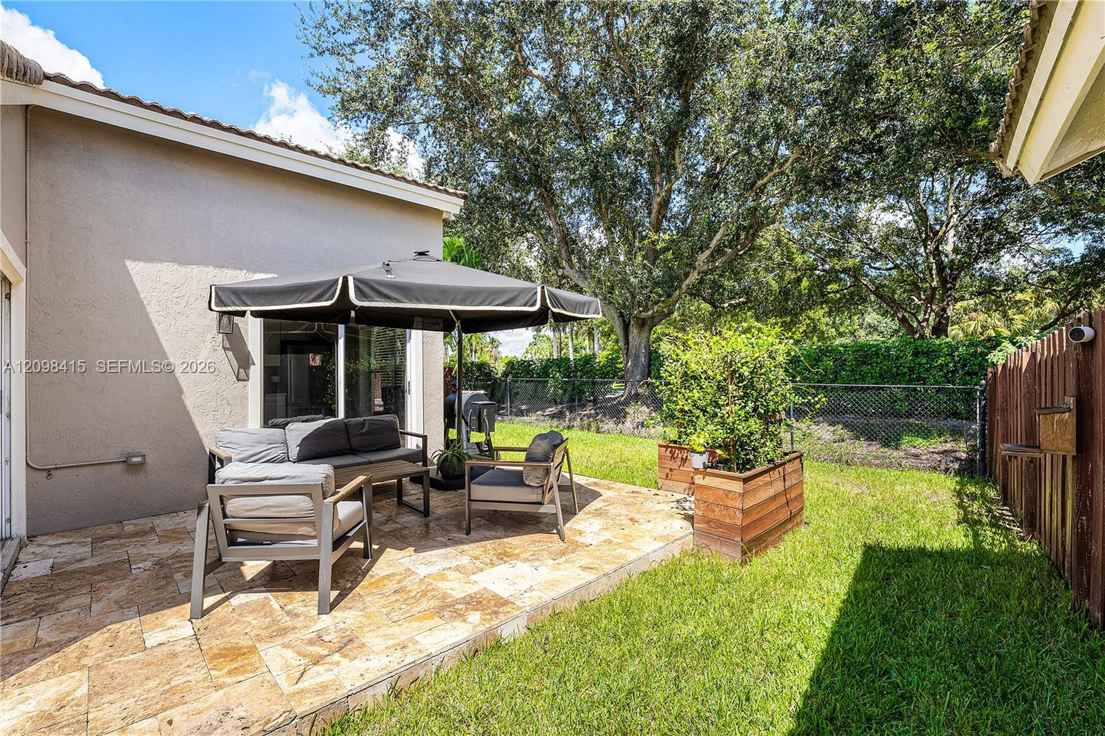 Backyard patio with cushioned seating beneath an umbrella beside raised planters and a fenced lawn.