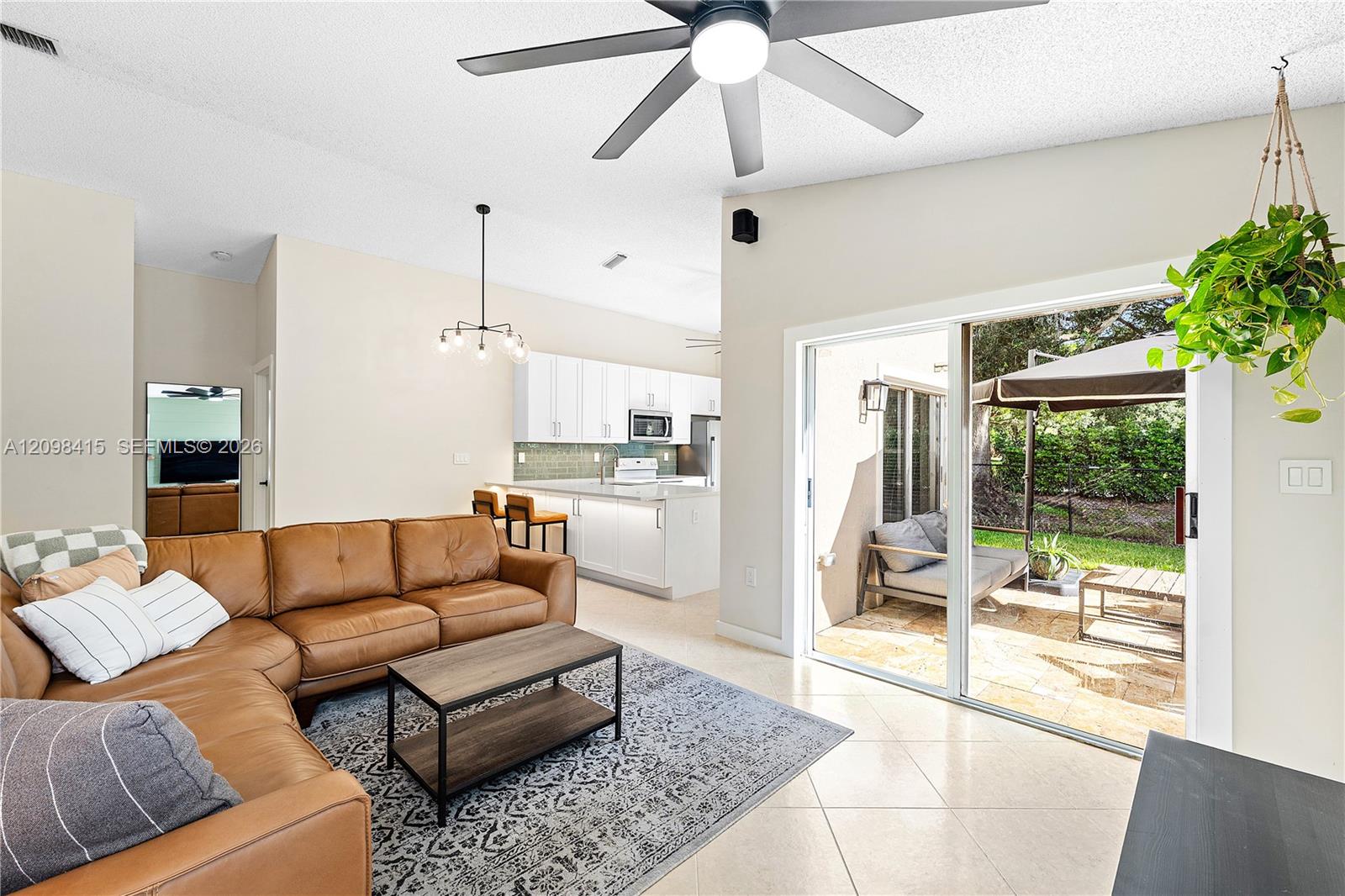 Furnished living room with a tan leather sectional, open kitchen, and sliding doors to a sunny patio.