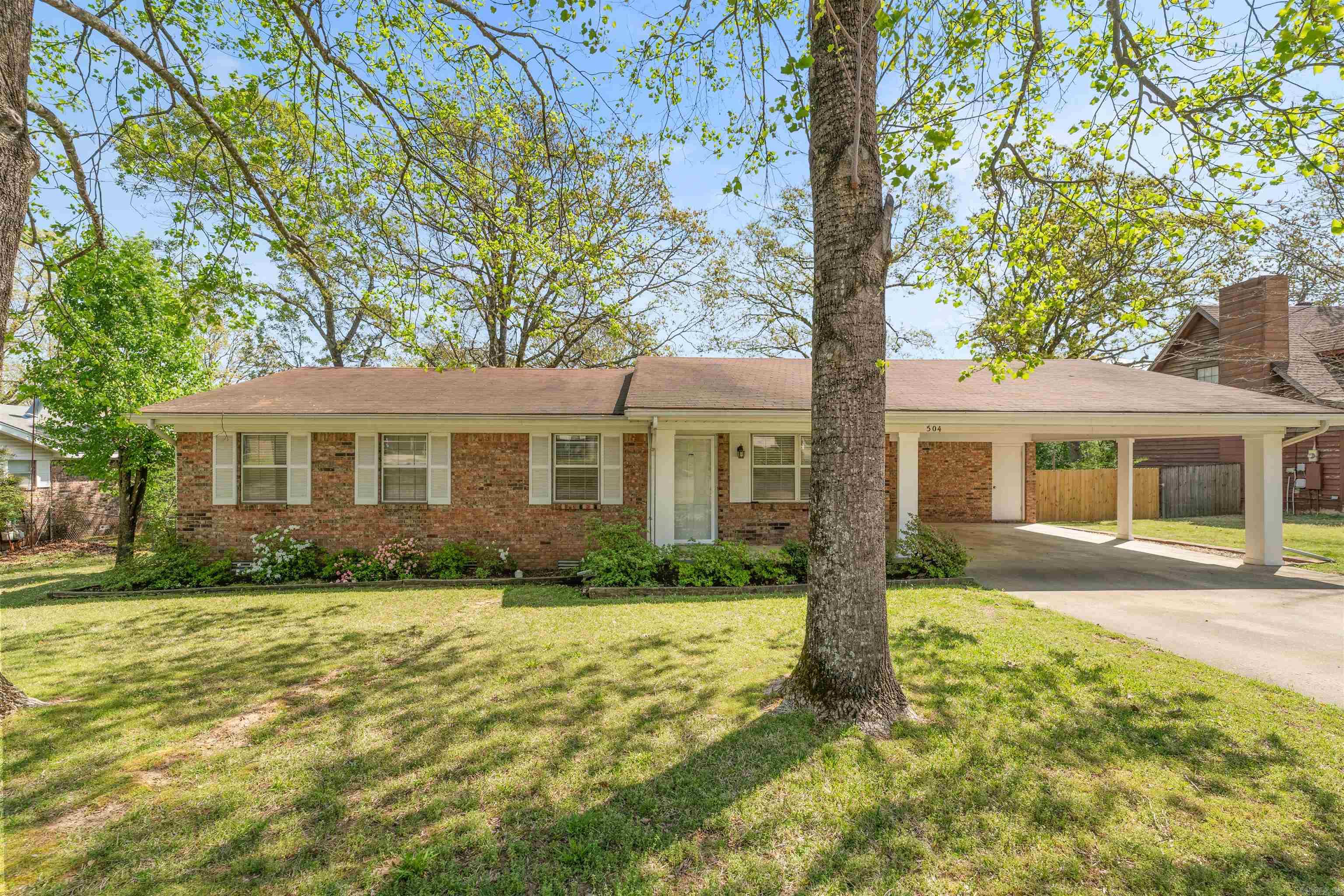 Brick ranch house with an attached carport, mature trees, and a front lawn.