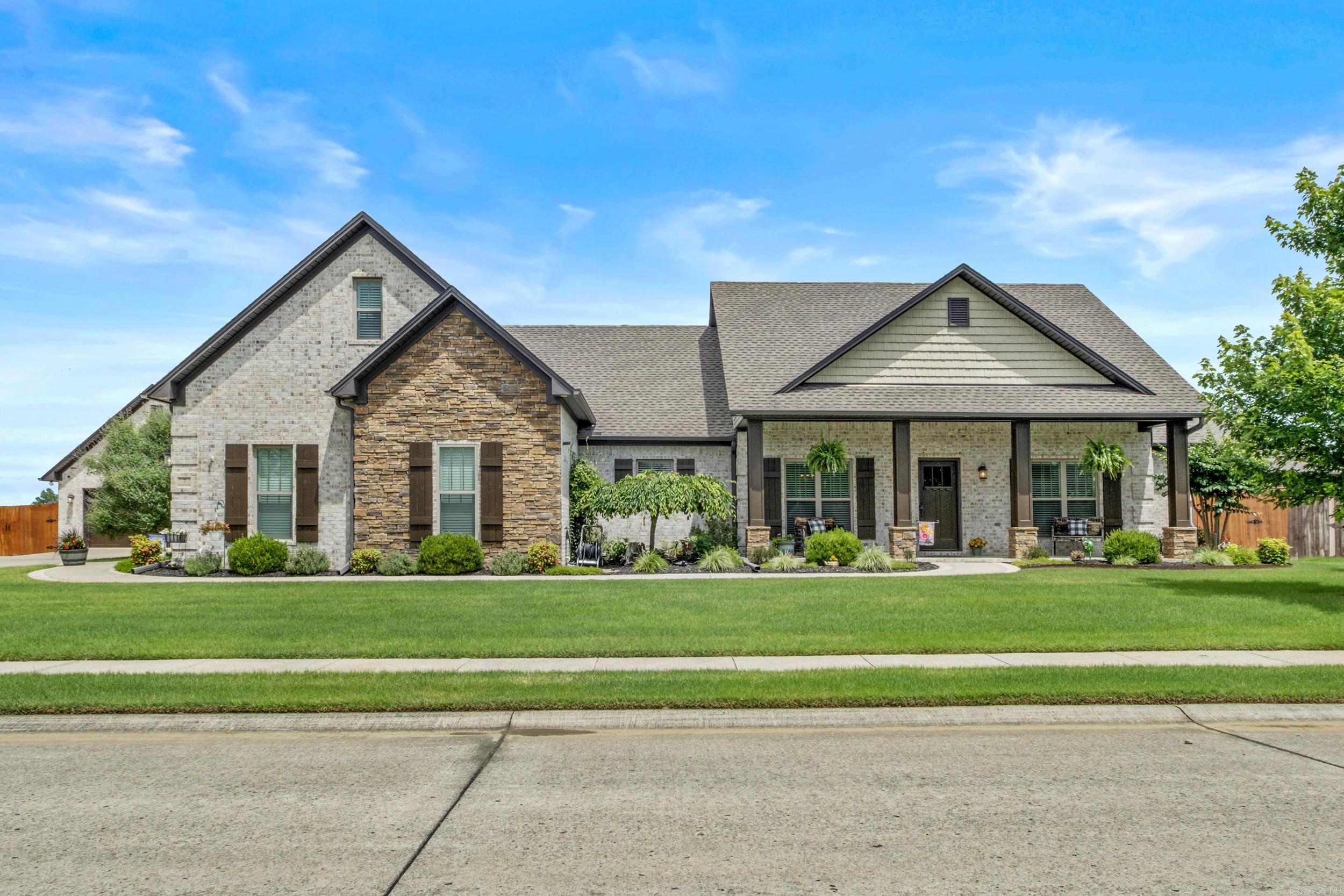 Gabled brick and stone house with a covered porch, landscaped garden beds, and a broad green lawn.