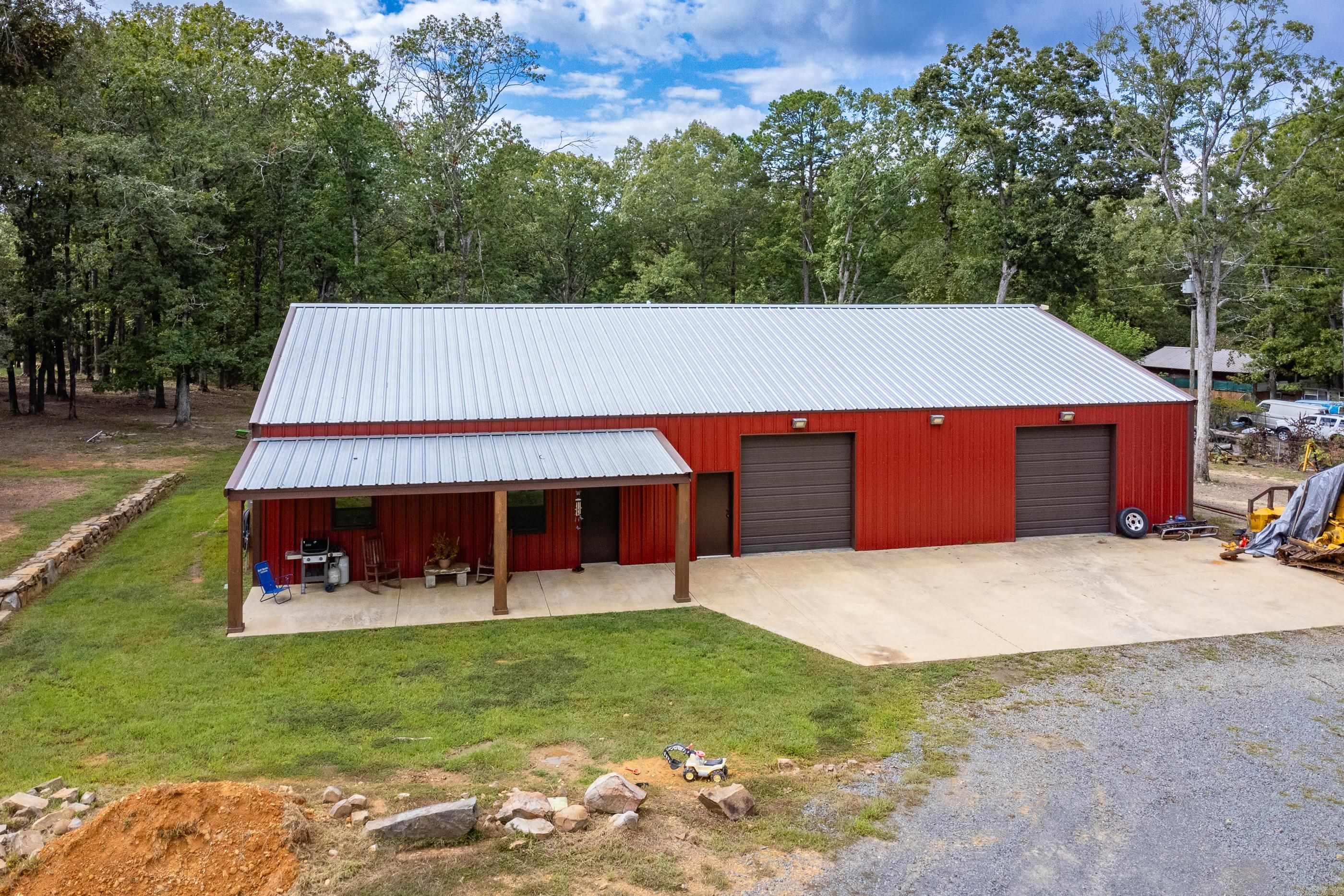 Red metal workshop with two garage doors and a covered porch, surrounded by trees.