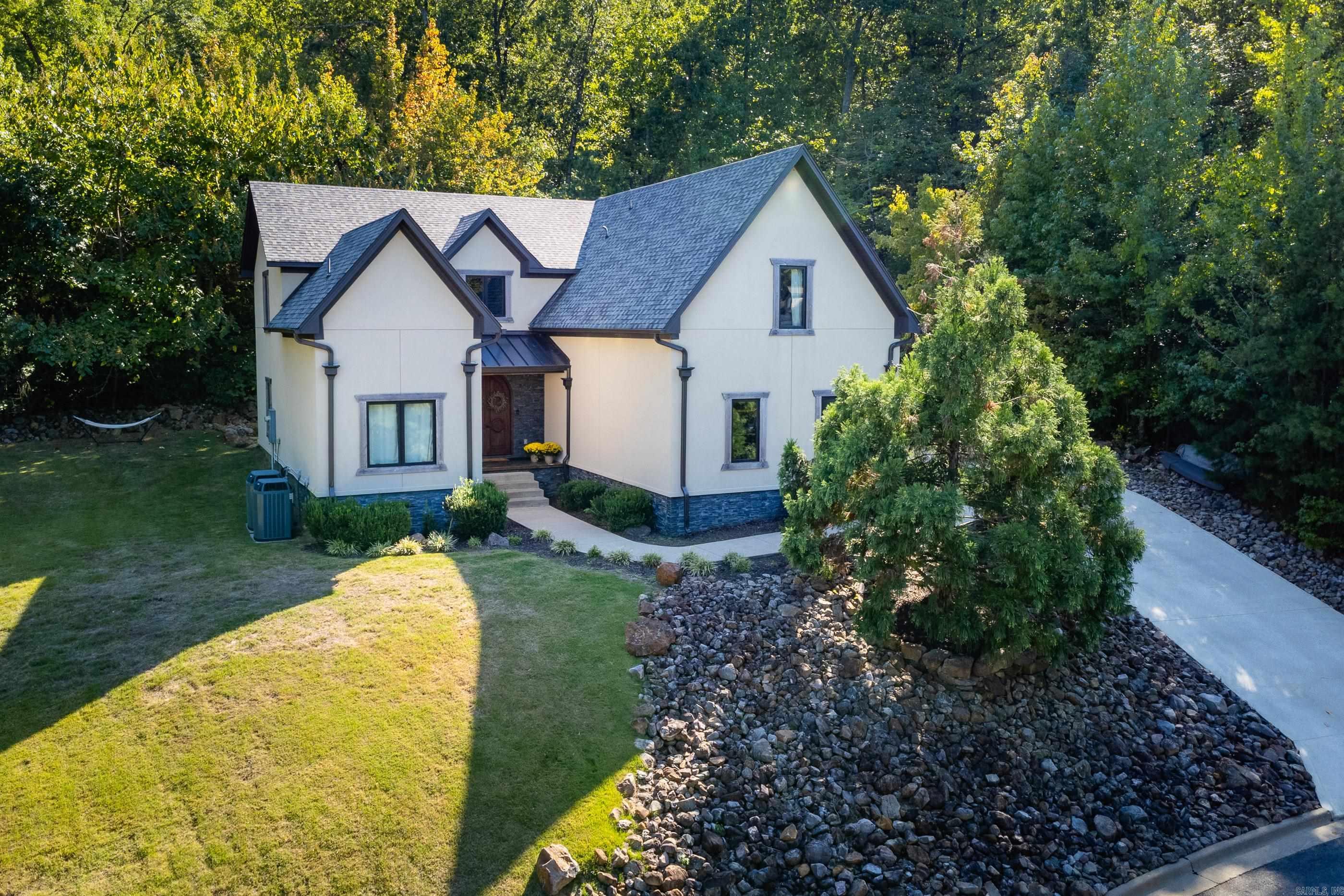 White two-story house with dark gabled roofs, landscaped rock beds, and a wooded backdrop.