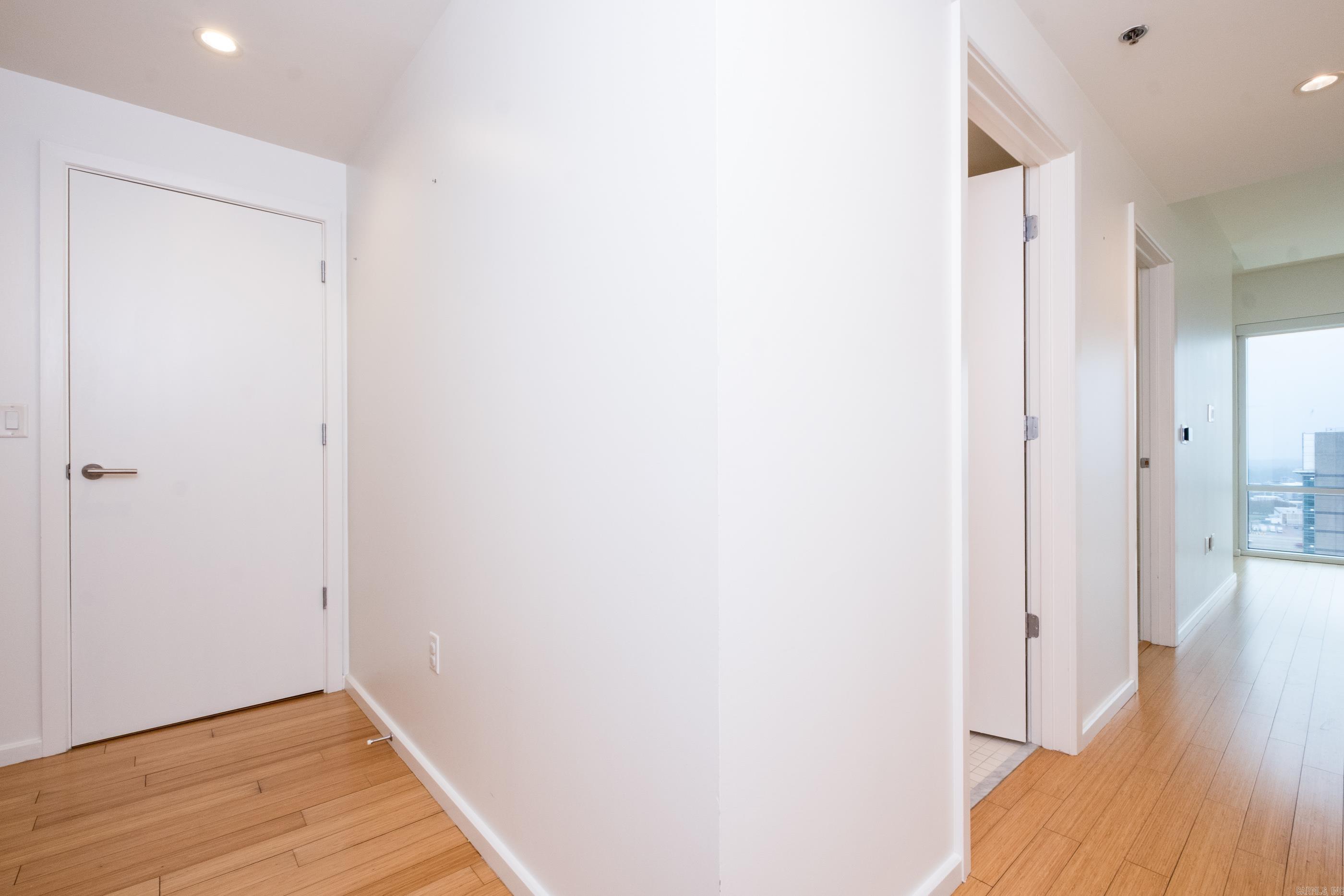 Empty apartment hallway with wood flooring, open doorways, and a large window at the far end.