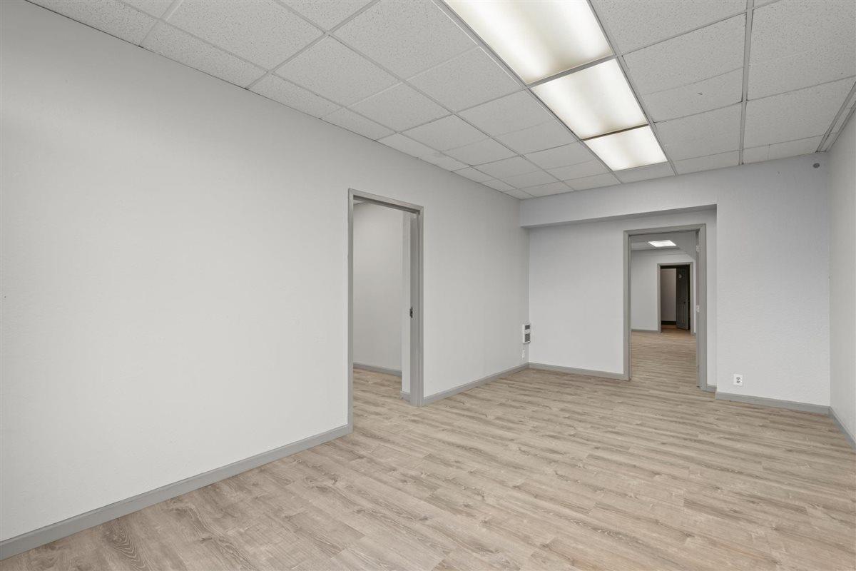 Vacant office room with wood-look flooring, fluorescent ceiling panels, and doorways to adjoining rooms.