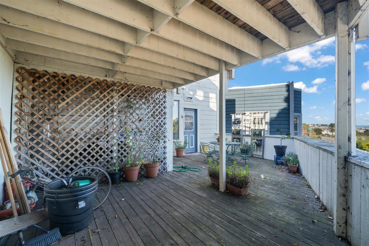 Covered wooden deck with lattice screening, potted plants, outdoor seating, and a view of a neighboring building.