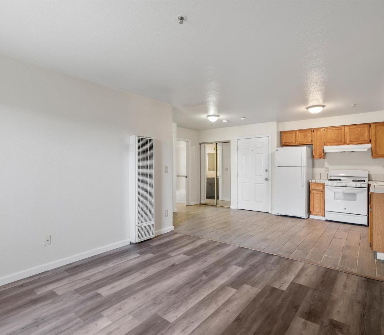 Open apartment living area and kitchen with wood cabinets, white appliances, and contrasting floors.