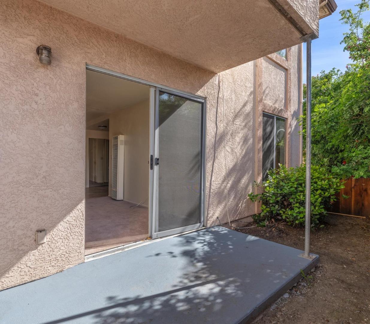 Covered concrete patio with an open sliding glass door and leafy hedge beside the stucco home.