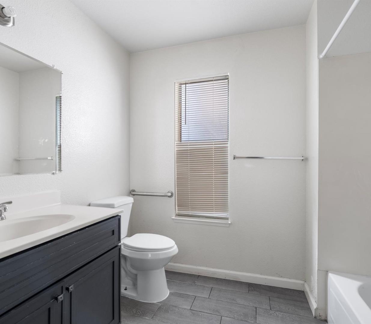 Bathroom with a dark vanity, toilet, window blinds, grab bars, and gray tile floor.