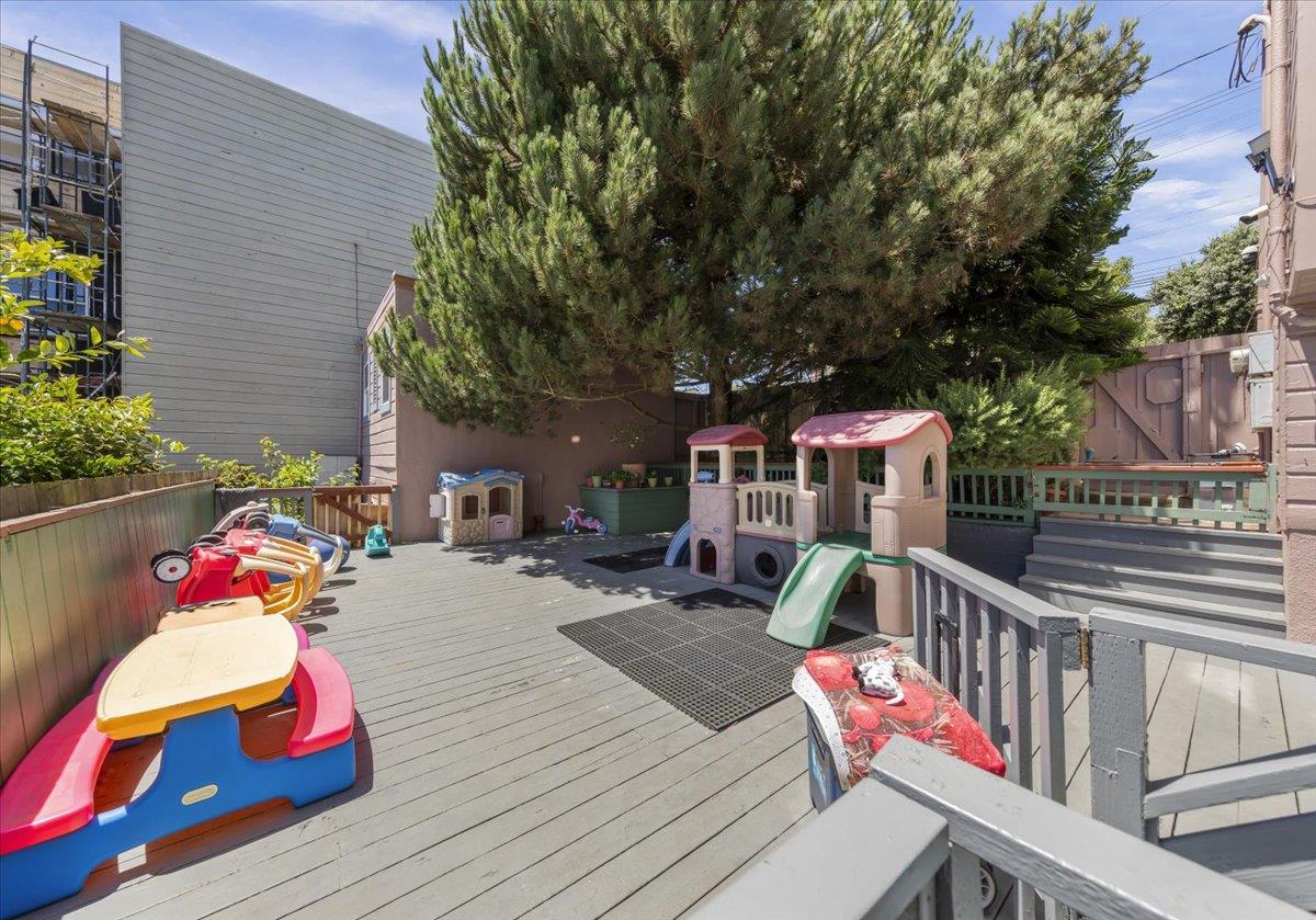 Outdoor deck play area with children’s play structures and a slide beneath a large pine tree.