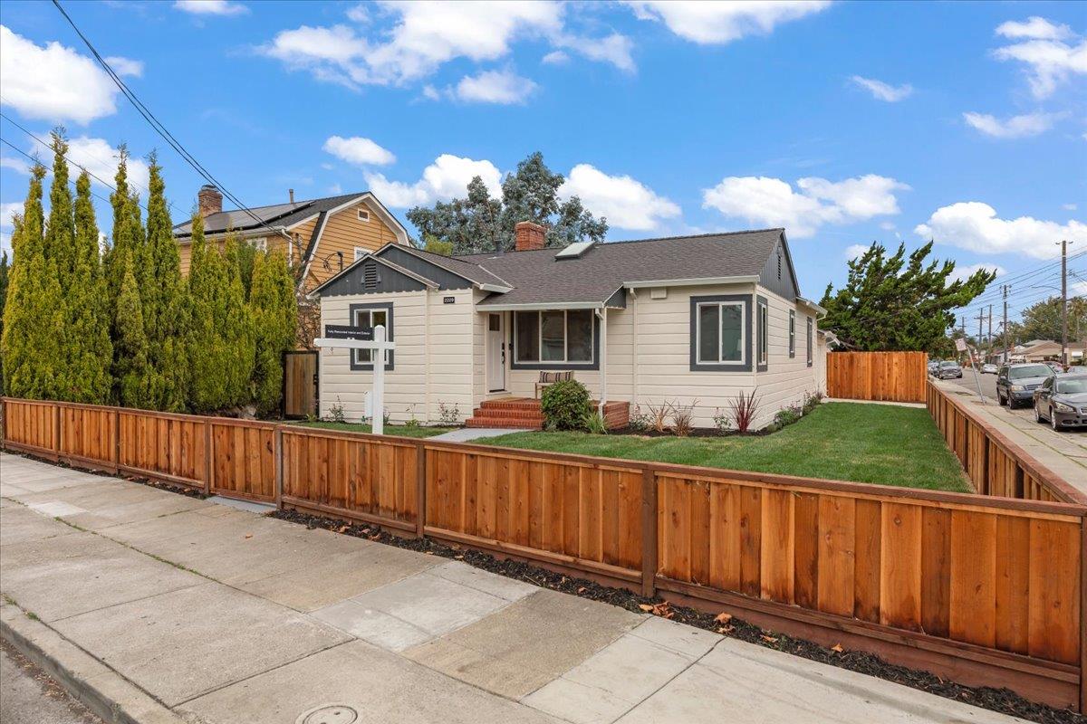Single-story beige house with a small porch, green lawn, and wooden fencing along the sidewalk.
