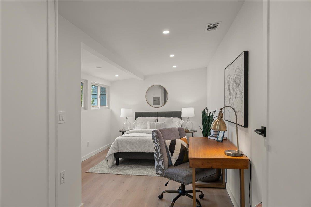 Staged bedroom with a neatly made bed, round mirror, and desk beside an upholstered chair.