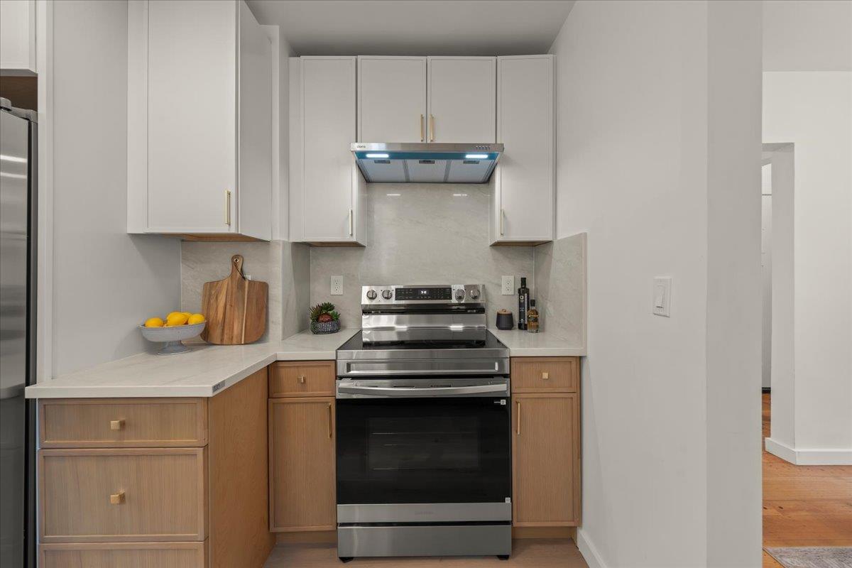 Kitchen with white cabinets, warm wood lower cabinetry, pale stone counters, and a stainless steel range.
