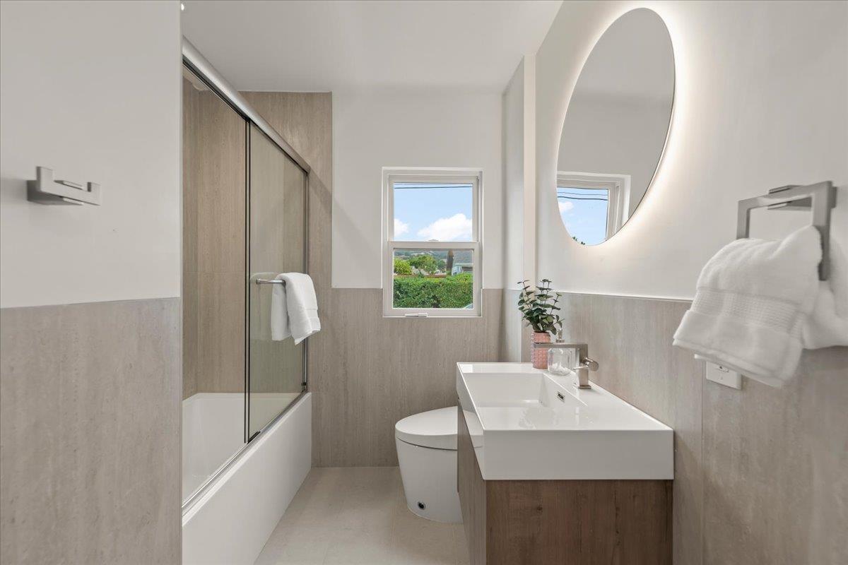 Updated bathroom with glass-enclosed tub, wood-look vanity, oval backlit mirror, and window overlooking greenery.
